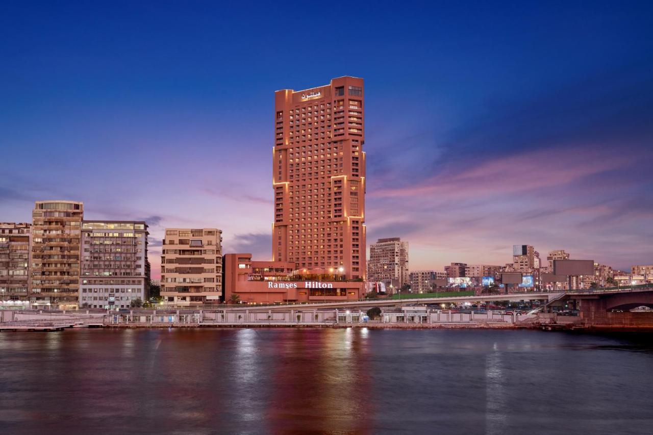 Ramses Hilton Hotel & Casino Kairo Exteriör bild The photo features a skyline view of a modern tall building, prominently lit against a twilight sky. The building is situated along a riverbank, showcasing a sleek architectural design. The surrounding area includes other low-rise buildings and struc
