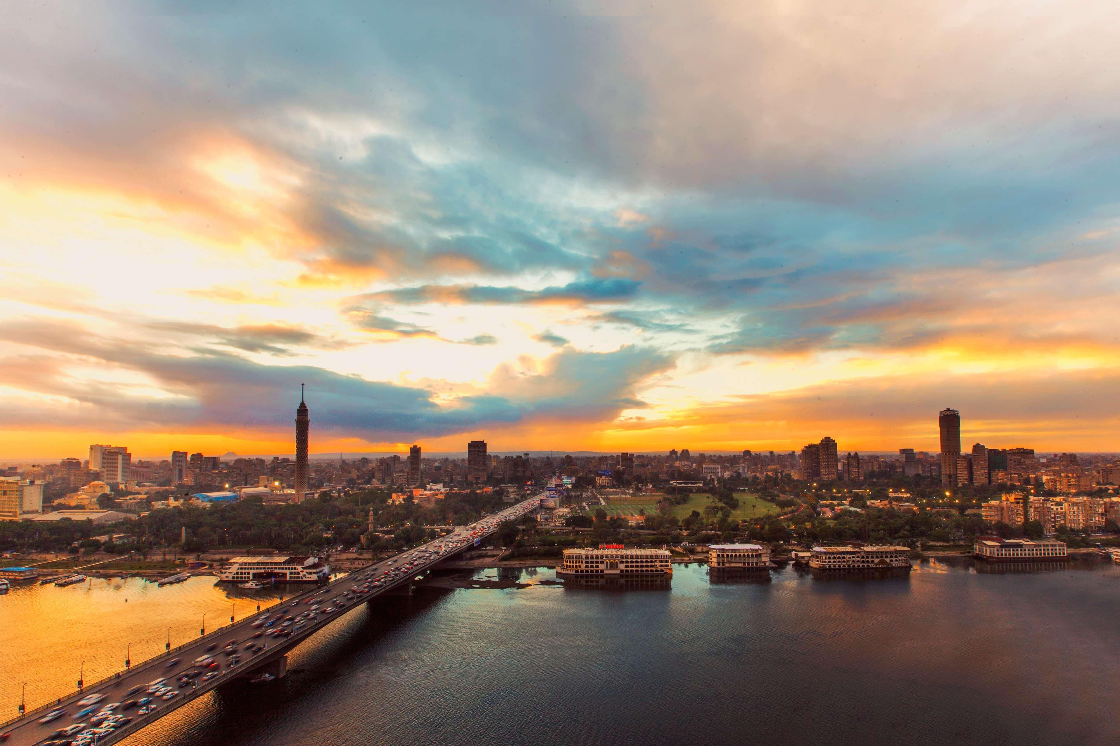Ramses Hilton Hotel & Casino Kairo Exteriör bild The photo captures a scenic view of a cityscape during sunset. In the foreground, there is a river reflecting the warm colors of the sky, which features shades of orange, blue, and purple. A bridge spans the river, connecting different parts of the c