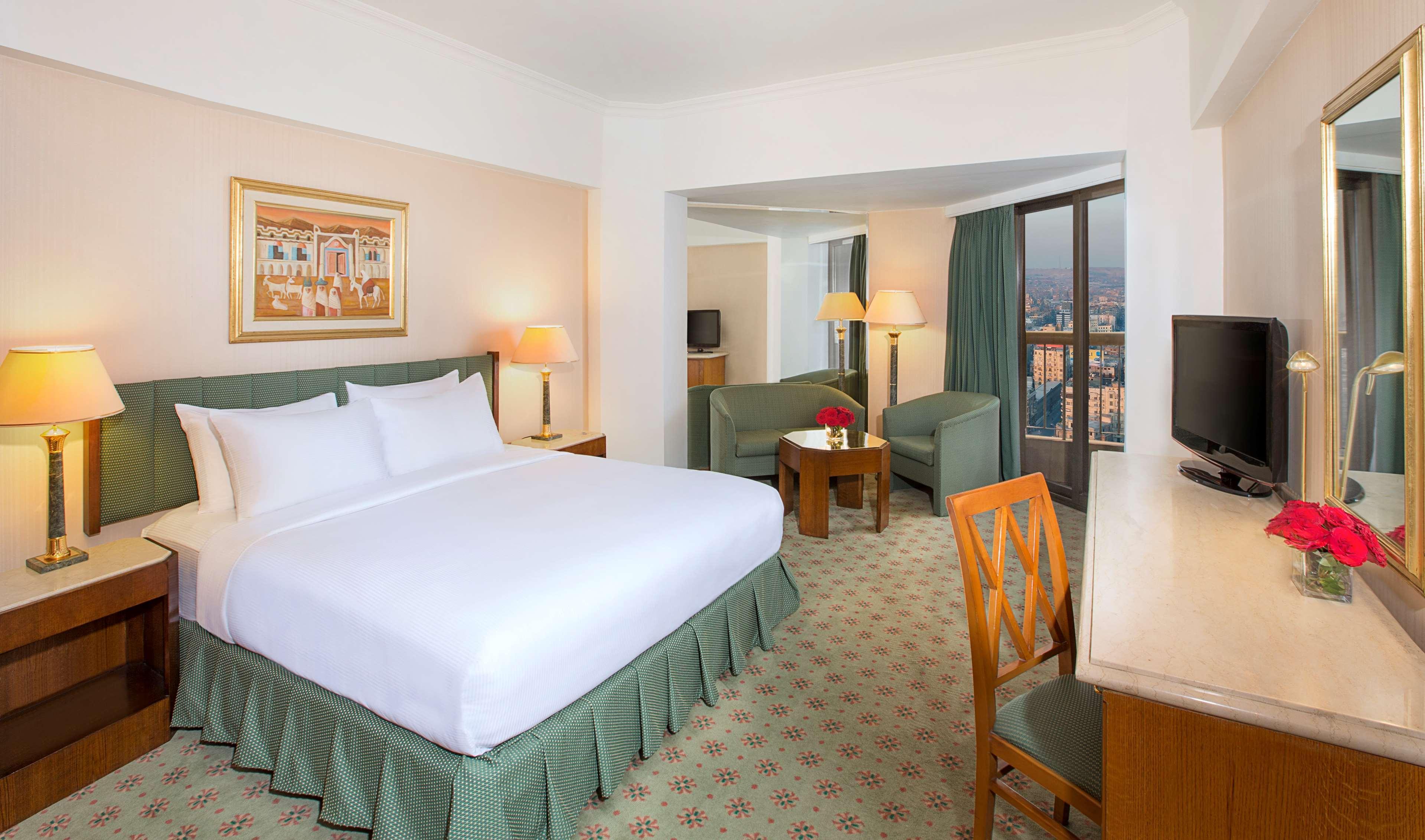 Ramses Hilton Hotel & Casino Kairo Exteriör bild The photo shows a hotel room that features a king-size bed with a white bedspread and a green skirt. There is a piece of artwork hanging on the wall above the bed. To one side, there are two lamps providing light, and a seating area is visible with t
