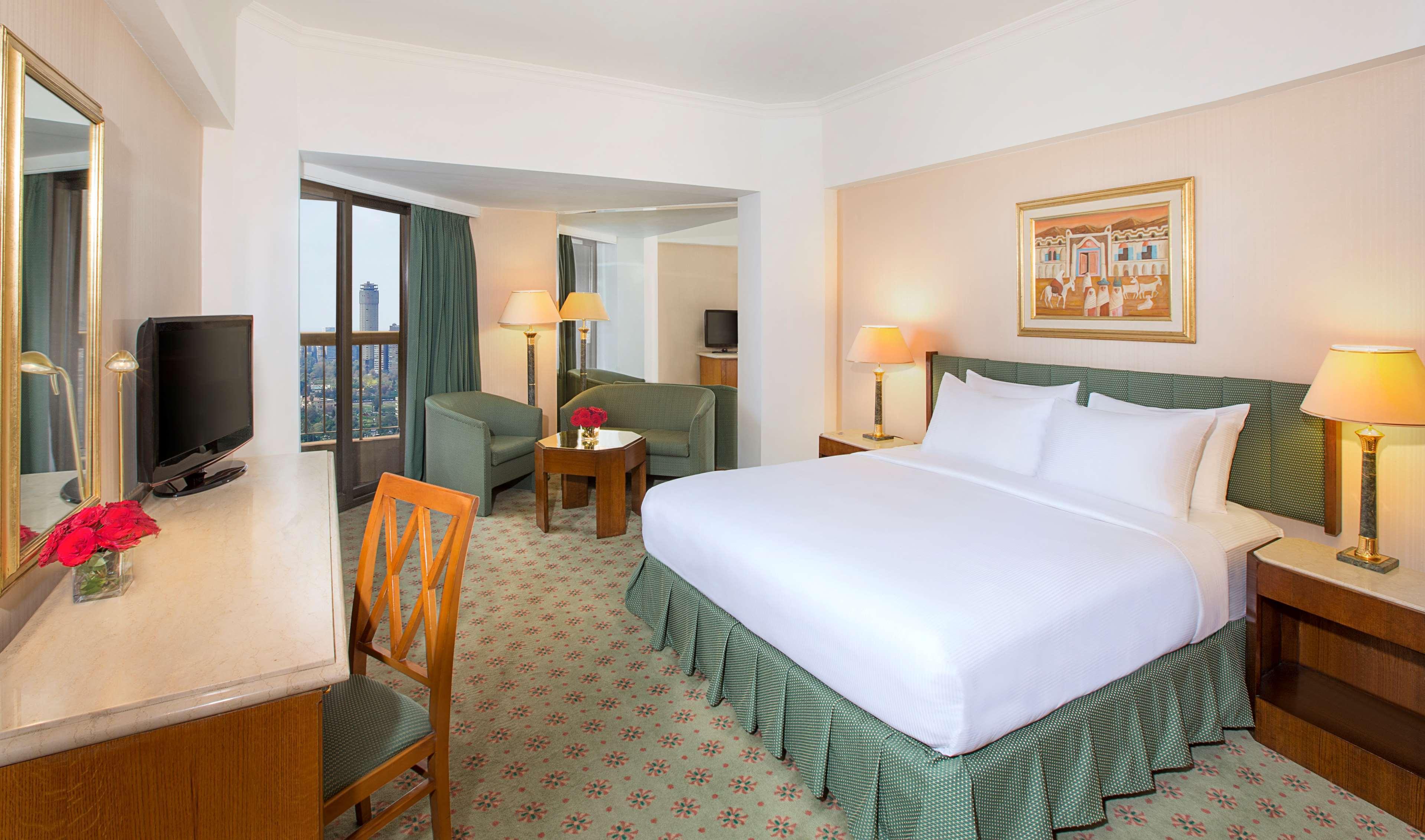 Ramses Hilton Hotel & Casino Kairo Exteriör bild The photo shows a hotel room that features a large bed covered in white linens, which has a green skirt. There are two chairs with a small table in front of them, creating a cozy seating area. A television is mounted on the wall, and there are window