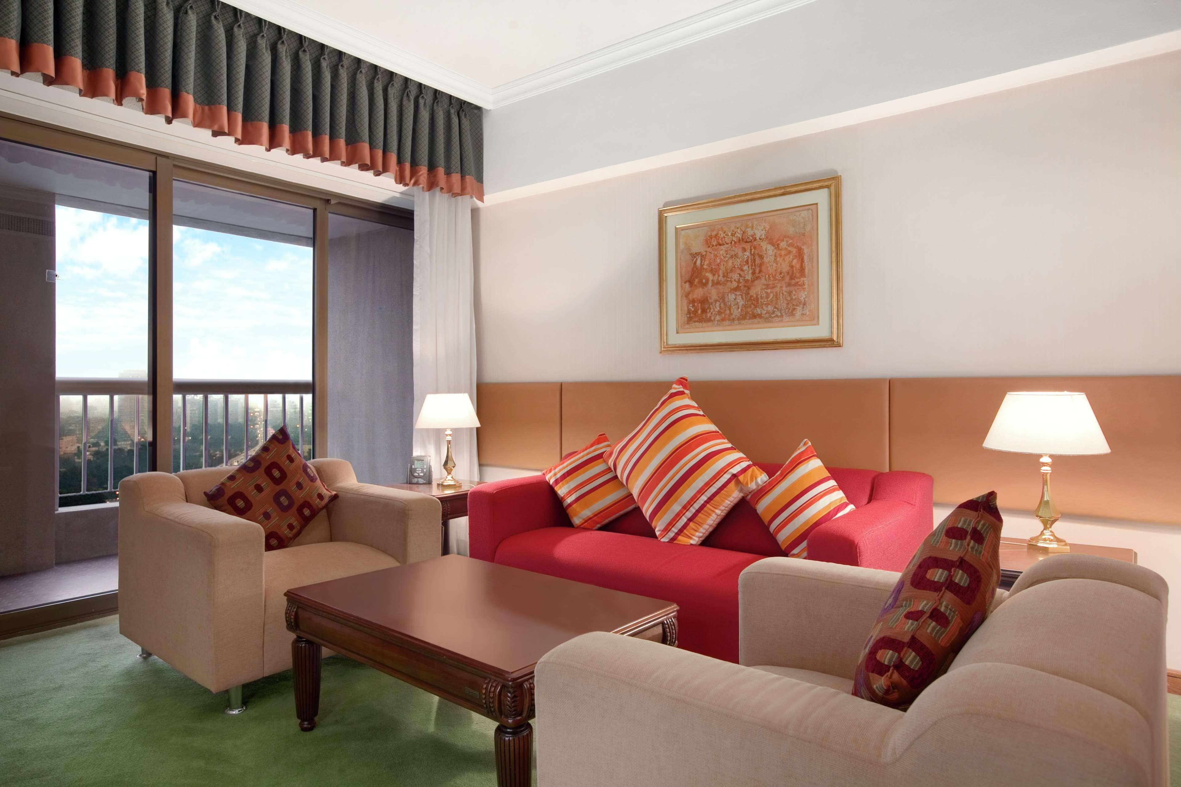Ramses Hilton Hotel & Casino Kairo Exteriör bild The photo depicts a cozy hotel room or living space. It features a red sofa adorned with striped cushions, complemented by two beige armchairs. In front of the seating area is a wooden coffee table. The room has a large window that allows natural lig