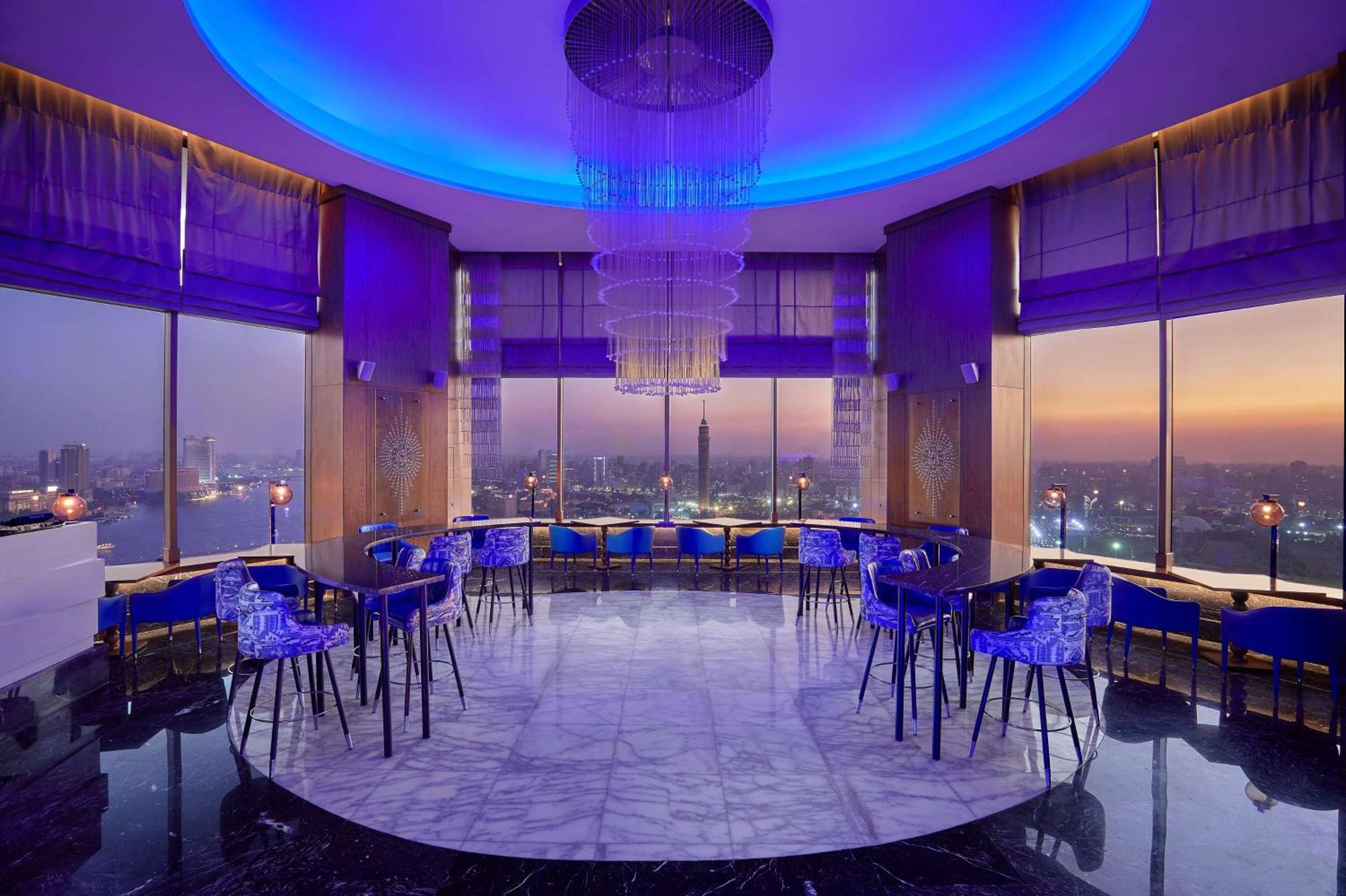 Ramses Hilton Hotel & Casino Kairo Exteriör bild The photo depicts a modern, elegantly designed lounge or bar area with large windows offering a panoramic view. The interior features stylish furniture, including transparent blue chairs arranged around black, round tables. The space is illuminated b