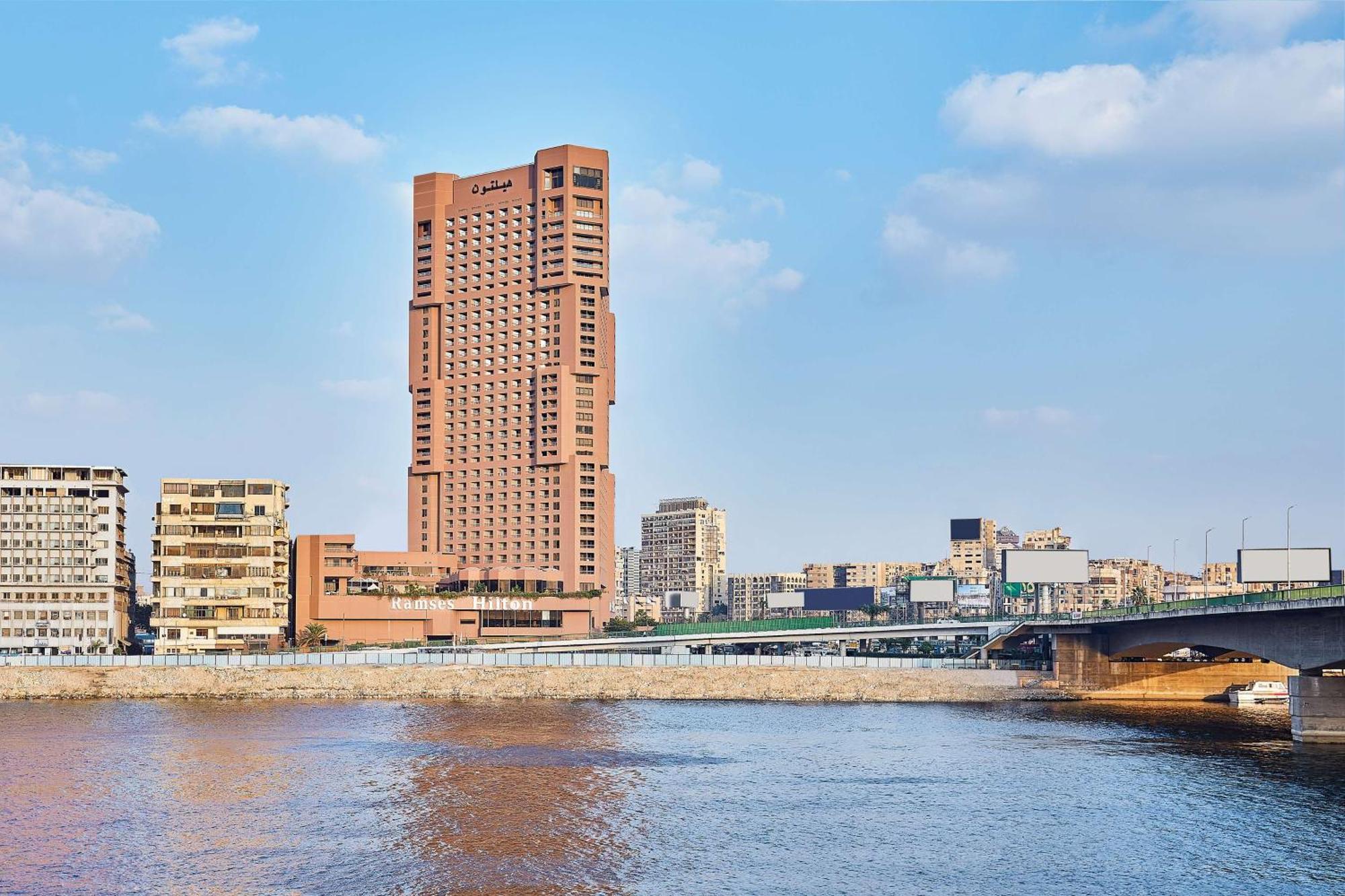 Ramses Hilton Hotel & Casino Kairo Exteriör bild The photo depicts a scene featuring a large, modern skyscraper with a distinctive architectural design, situated near a river. The building is prominently placed against a backdrop of a clear blue sky with scattered clouds. In the foreground, the wat