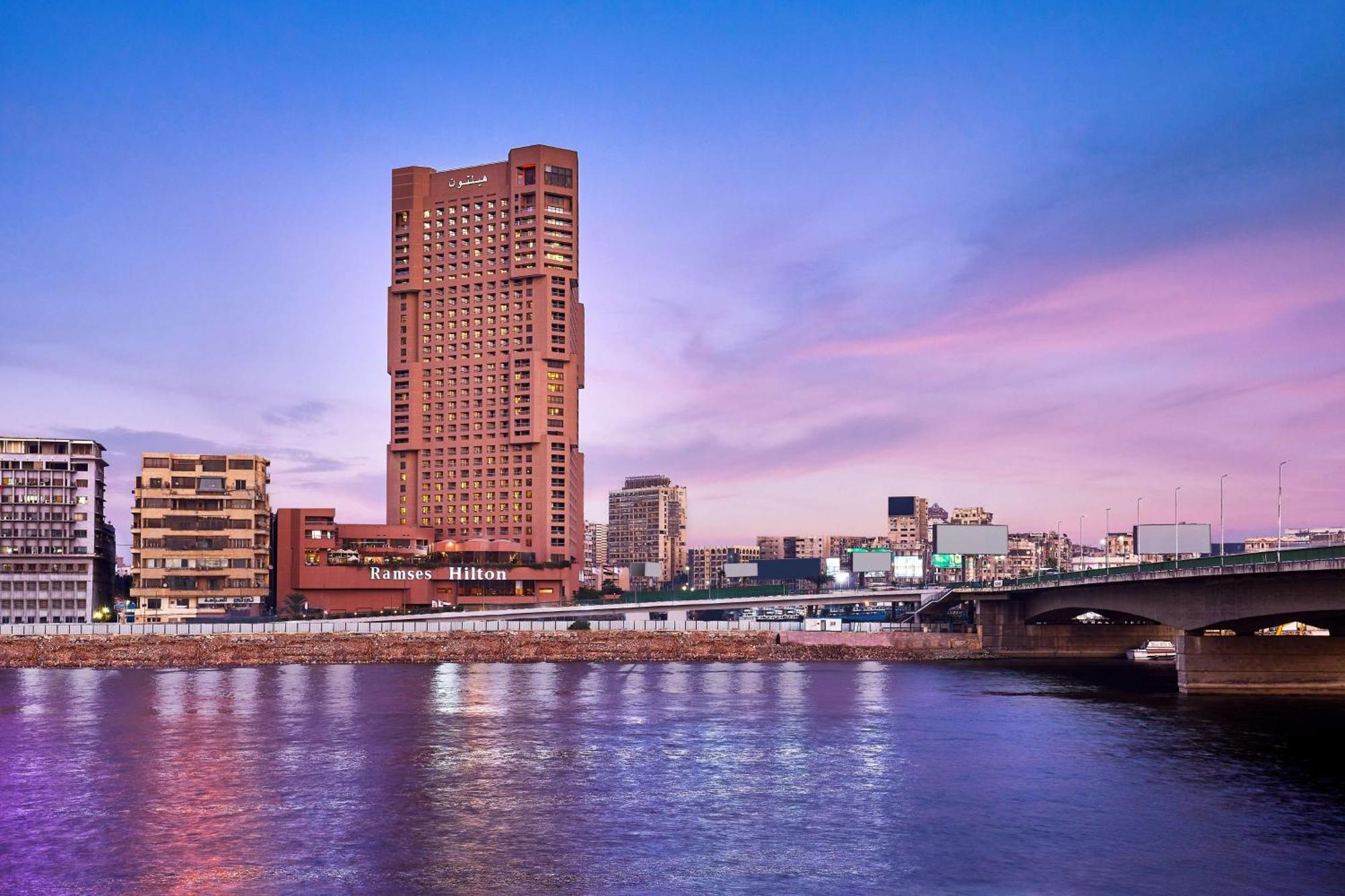 Ramses Hilton Hotel & Casino Kairo Exteriör bild The photo depicts a cityscape featuring a prominent tall building with a distinctive architectural design. It stands near a river, which reflects the colorful sky above, suggesting that it may be during sunset or sunrise. In the background, various o