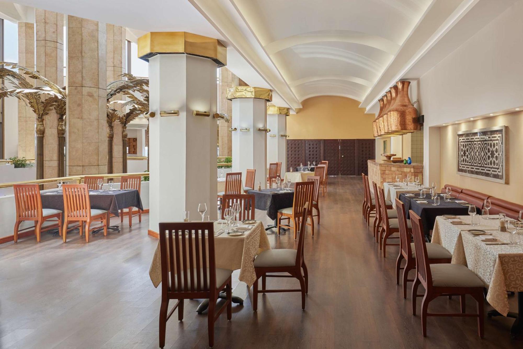 Ramses Hilton Hotel & Casino Kairo Exteriör bild The photo shows the interior of a restaurant. It features a modern design with light-colored walls and wooden flooring. There are several dining tables arranged with tablecloths and chairs made of wood. The space is well-lit, with large windows allow
