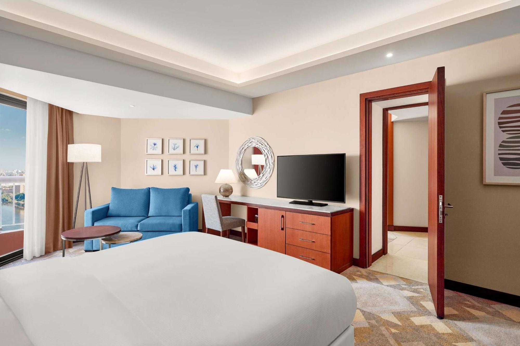 Ramses Hilton Hotel & Casino Kairo Exteriör bild The photo shows a modern hotel room. On one side, there is a large bed with white bedding. Opposite the bed, there is a seating area featuring a blue couch and a small coffee table. Next to the couch, there is a lamp providing light. A TV is mounted 